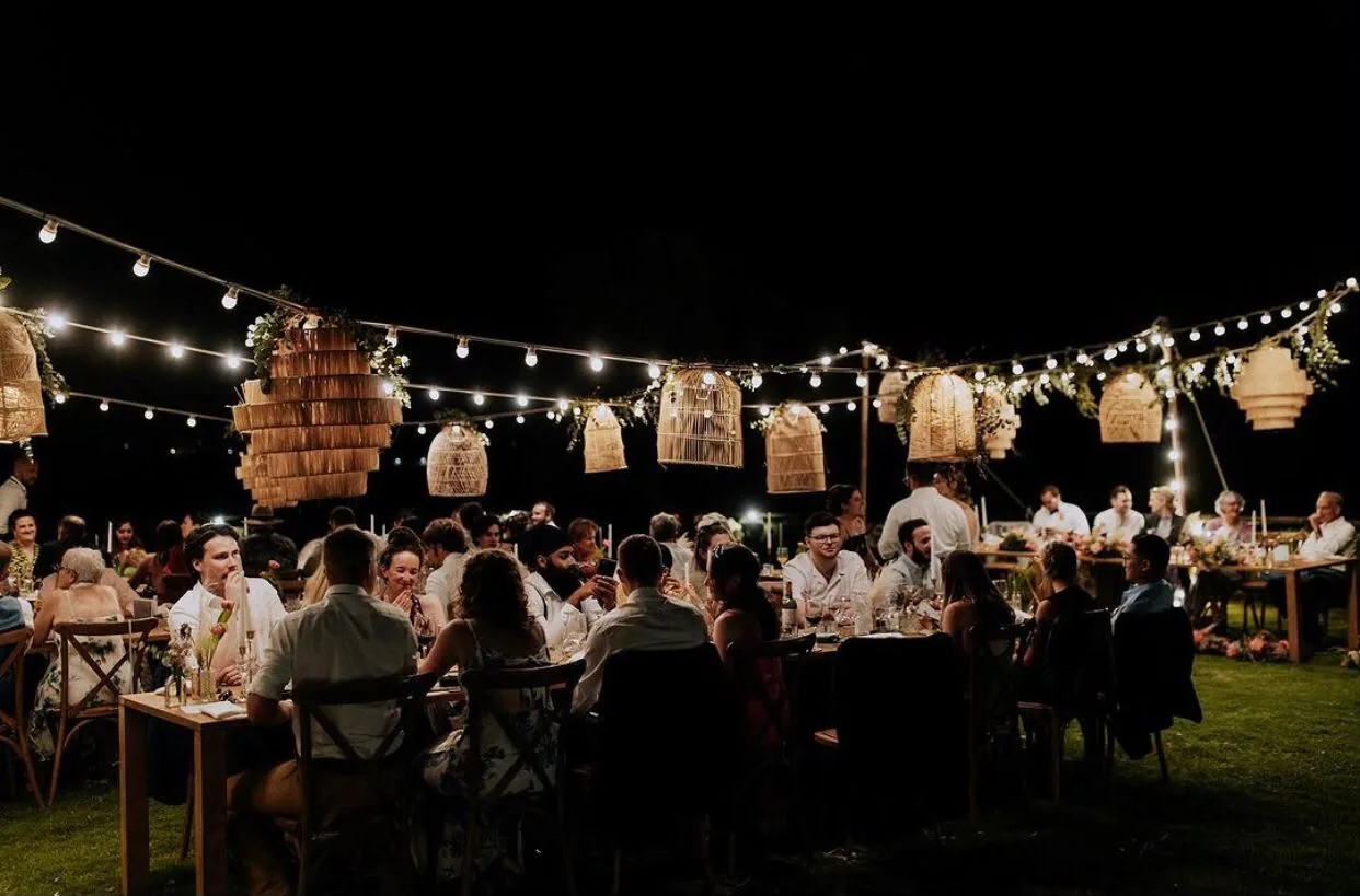 Cena al aire libre bajo guirnaldas de luces y lámparas de mimbre — recepción nocturna con invitados.
