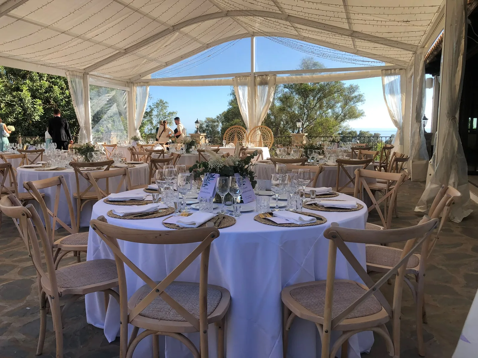 Carpa de boda con cortinas de lino y vistas al mar, mesas redondas vestidas de blanco bajo la estructura acristalada.