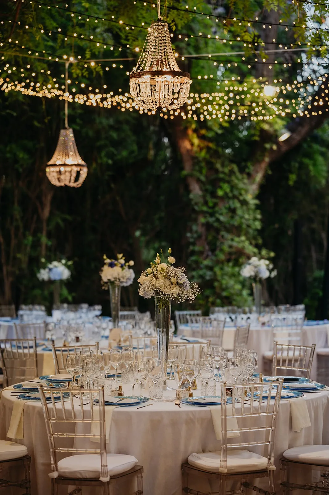 Candelabros colgantes entre los árboles sobre mesas redondas con centros florales, ambiente nocturno de boda
