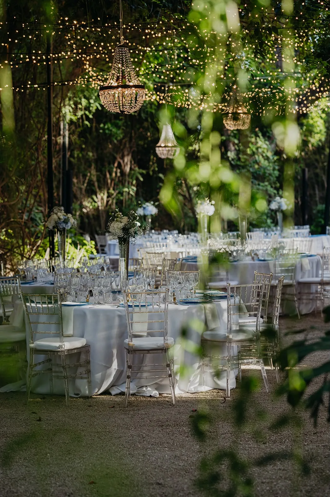 Candelabros entre el follaje con mesas largas al fondo, panorámica del jardín en boda al aire libre