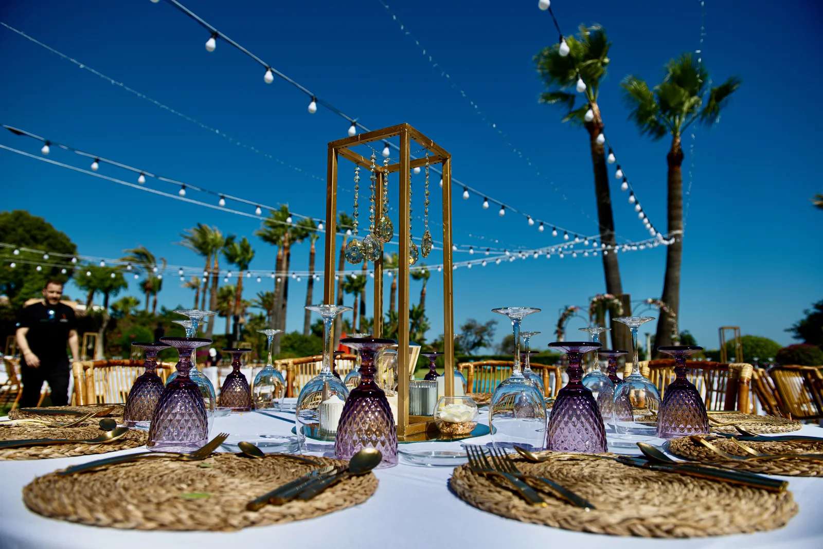 Premium outdoor wedding table detail: purple crystal glasses, gold geometric candle lantern, wicker placemats and gold cutlery with palm trees and blue sky