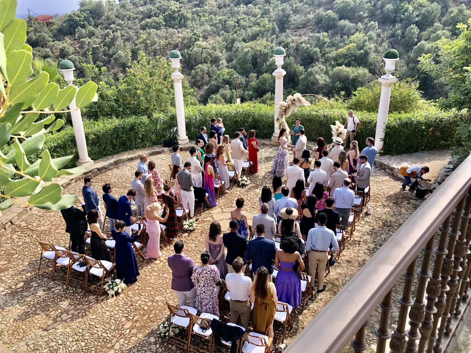 Aerial view of an outdoor wedding ceremony at a hillside venue: guests before the couple at a pampas grass arch flanked by white Classical columns