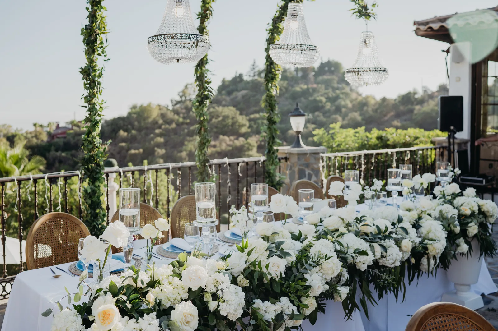 President table with abundant white roses and hydrangeas, crystal chandeliers on garland-wrapped columns, outdoor Jewish wedding event