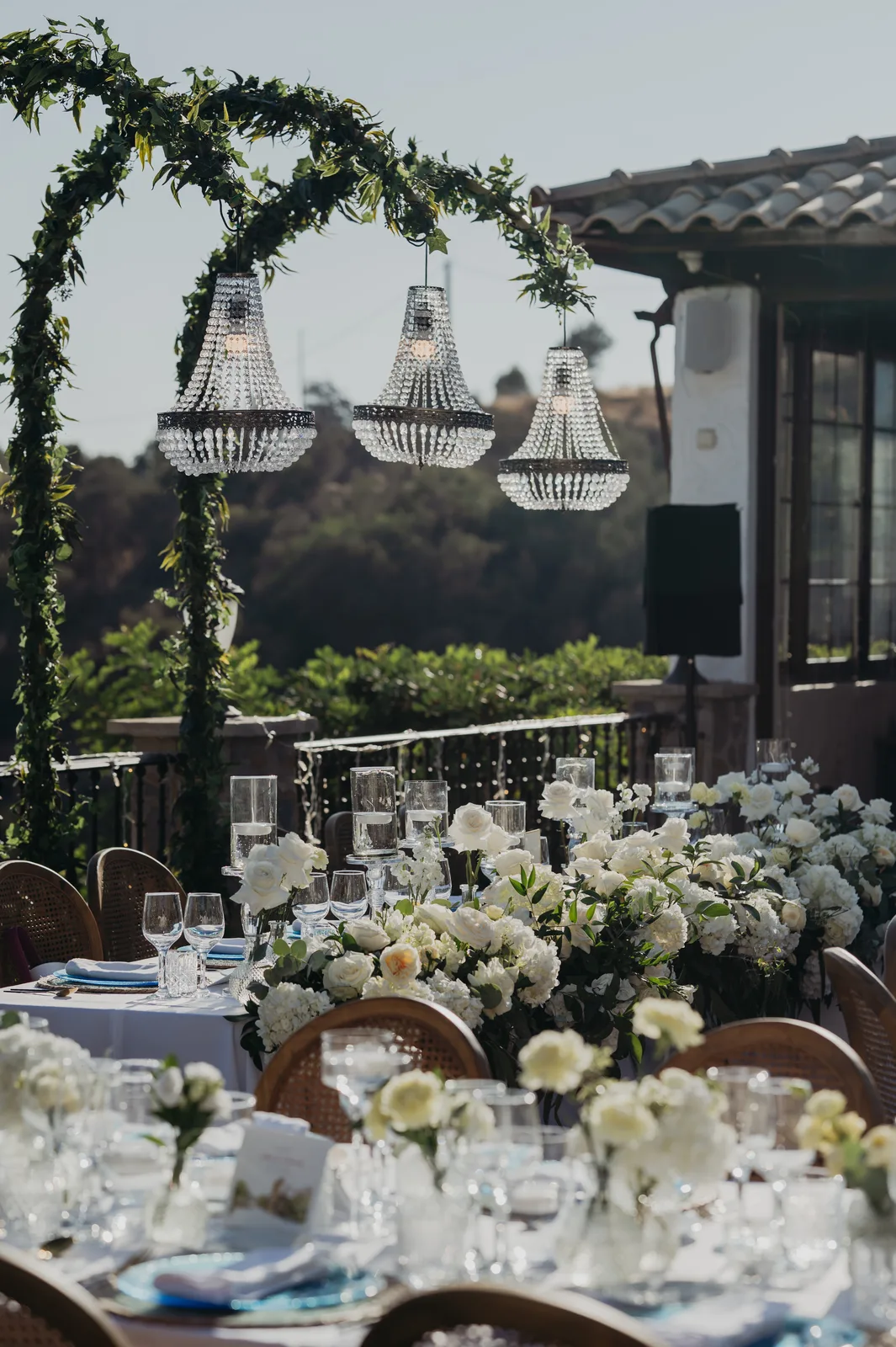 Outdoor terrace with crystal chandelier arch framed by lush foliage, long table with white rose runner and rattan chairs, Jewish wedding in Málaga