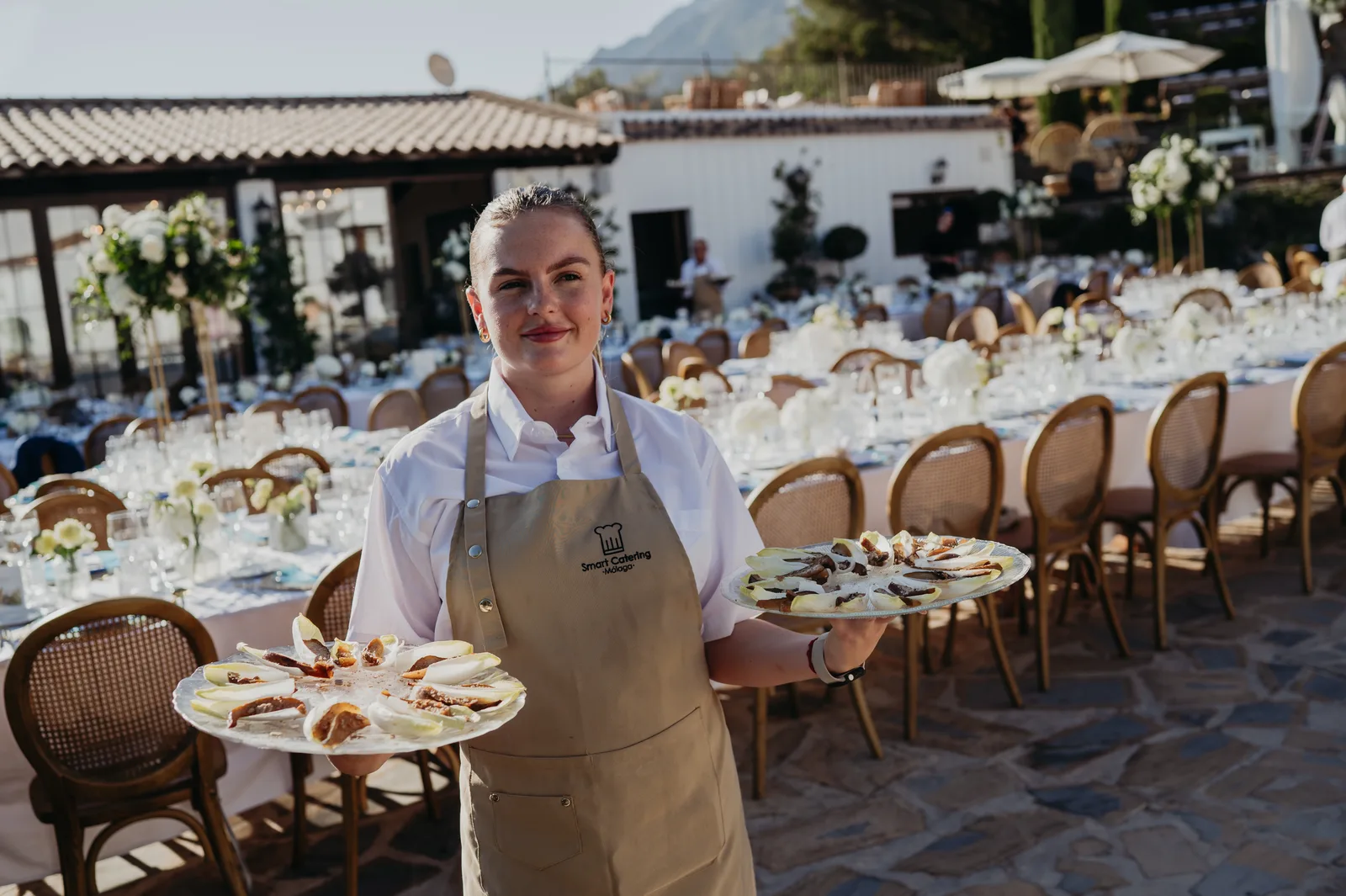 SmartCatering server carrying two starter plates with full kosher banquet — blue charger plates and white flower tables — in background