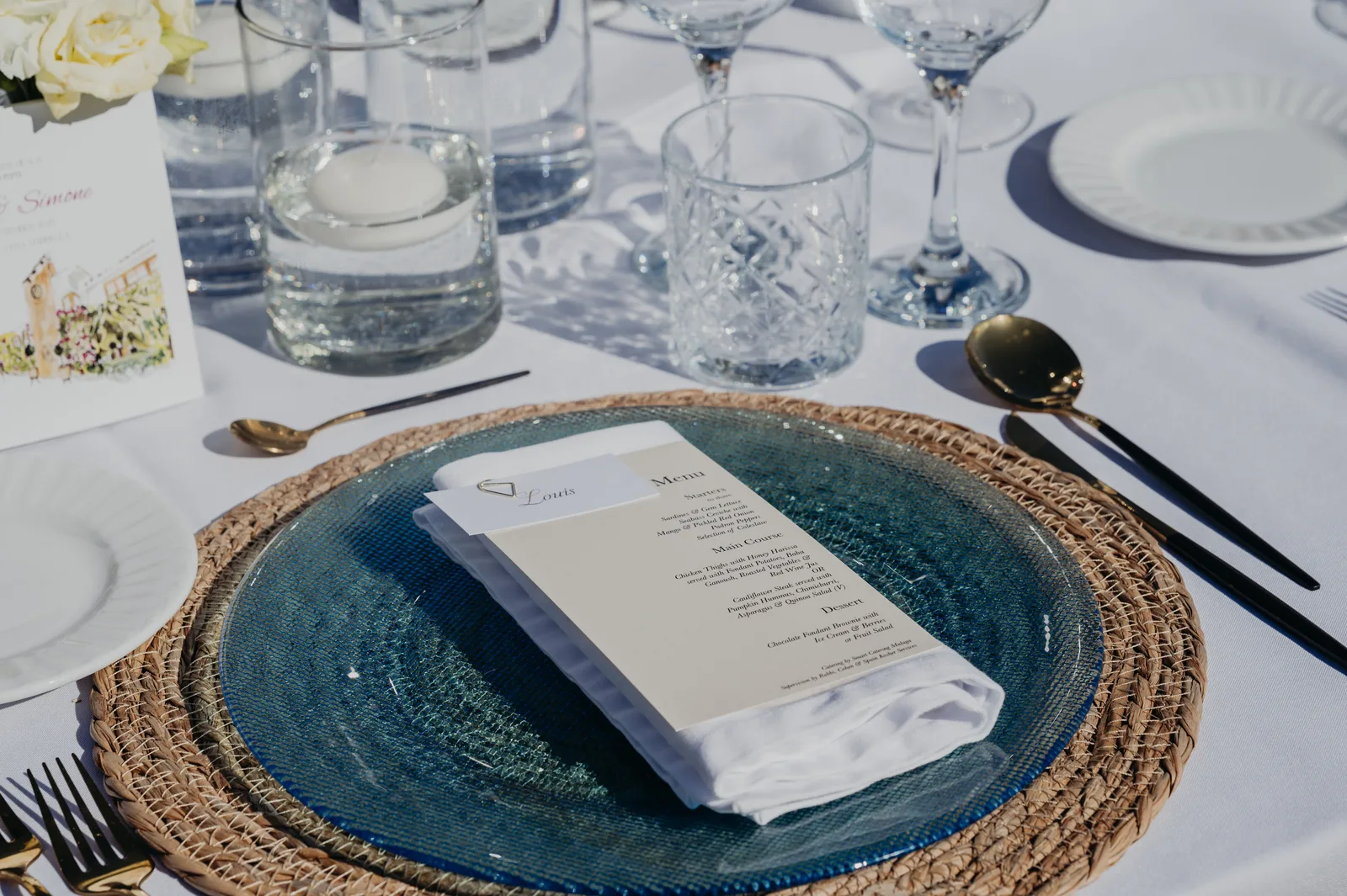 Kosher wedding table detail: blue glass charger on woven rattan mat, personalised menu card, floating candle, gold and black cutlery