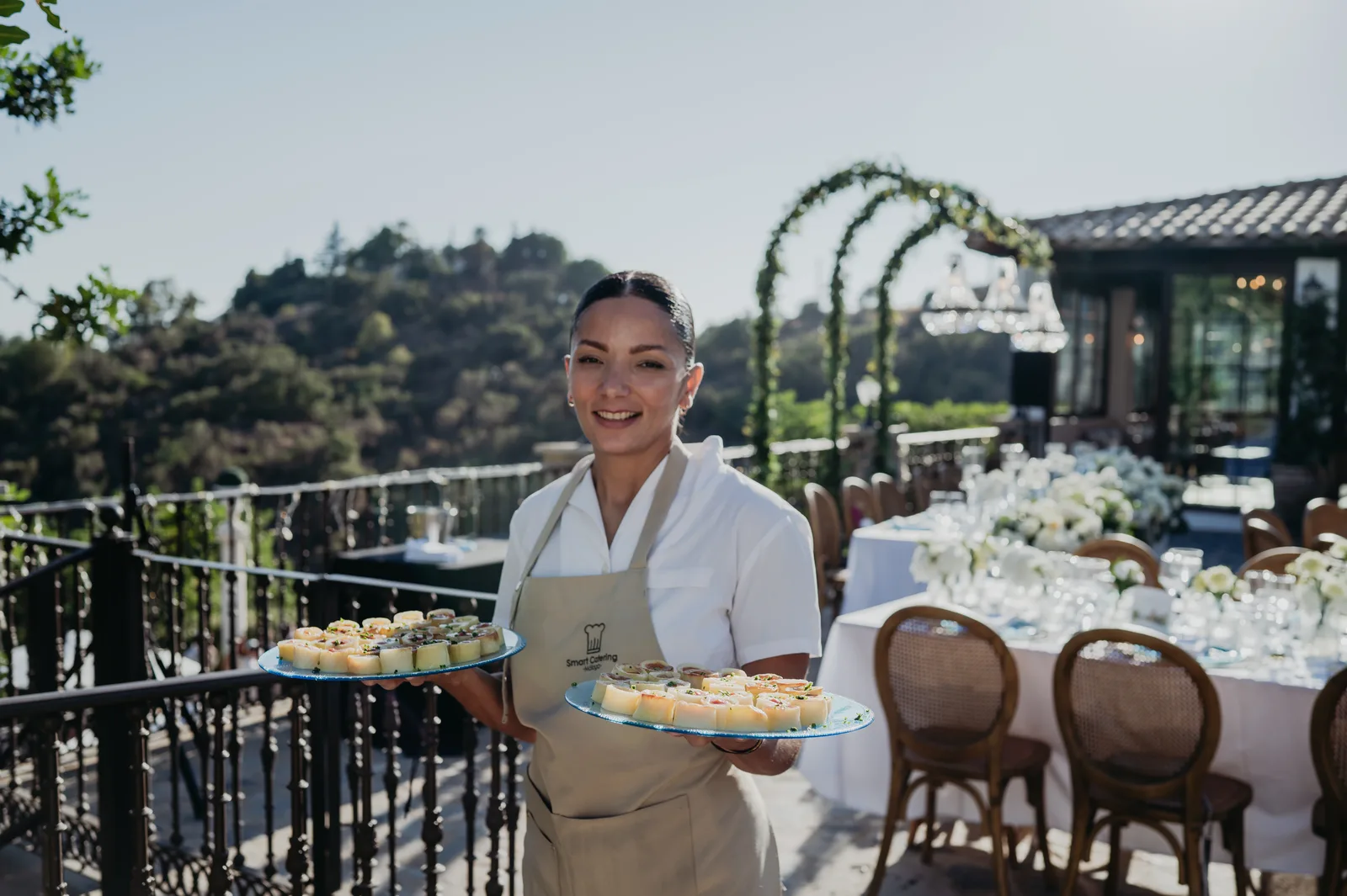 SmartCatering waitress carrying two trays of sushi-roll canapés at outdoor Jewish wedding terrace with white floral tables in background